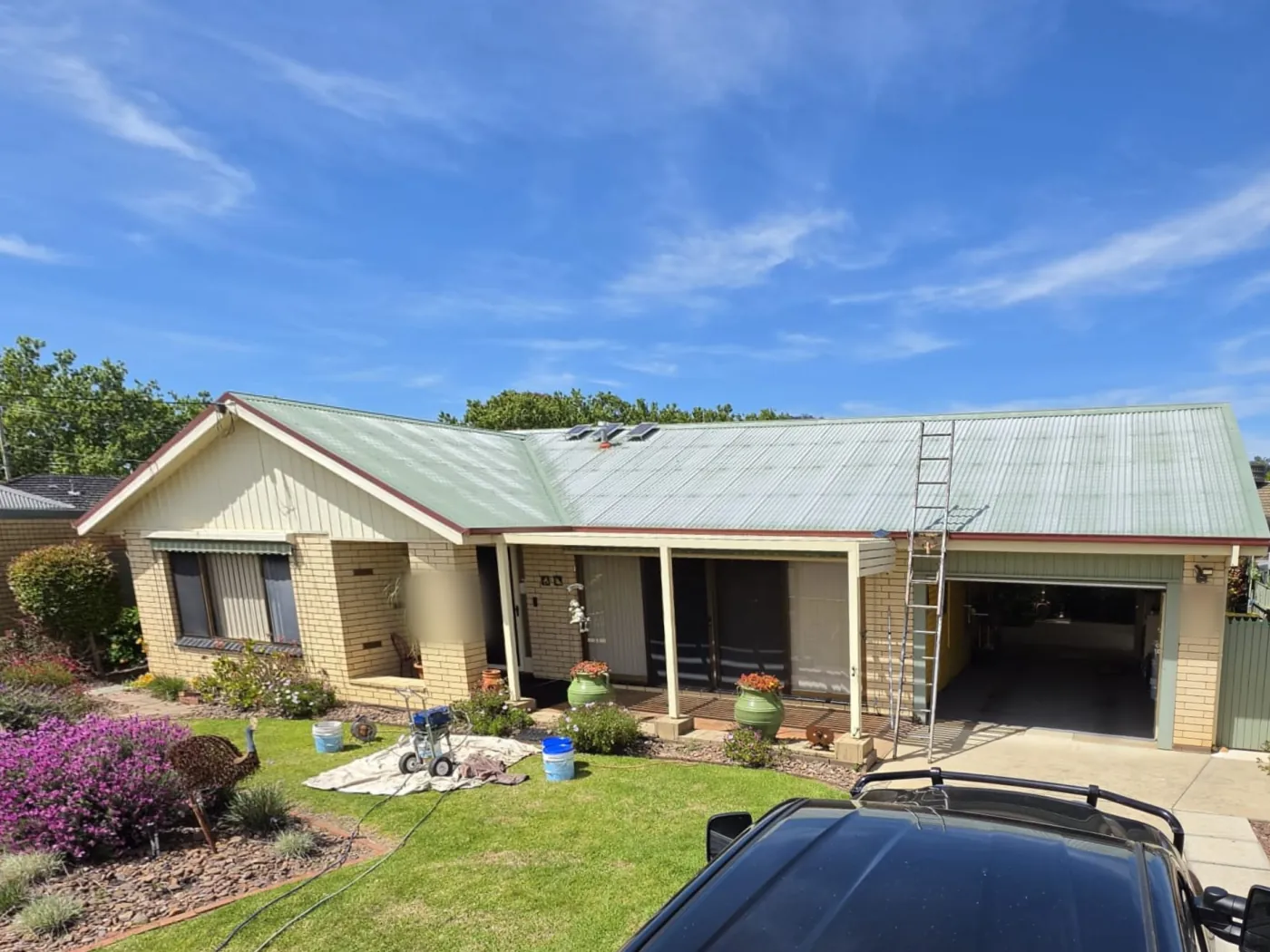 The same home from the same spot before, the old green paint on the roof faded and worn, with painting gear on the lawn